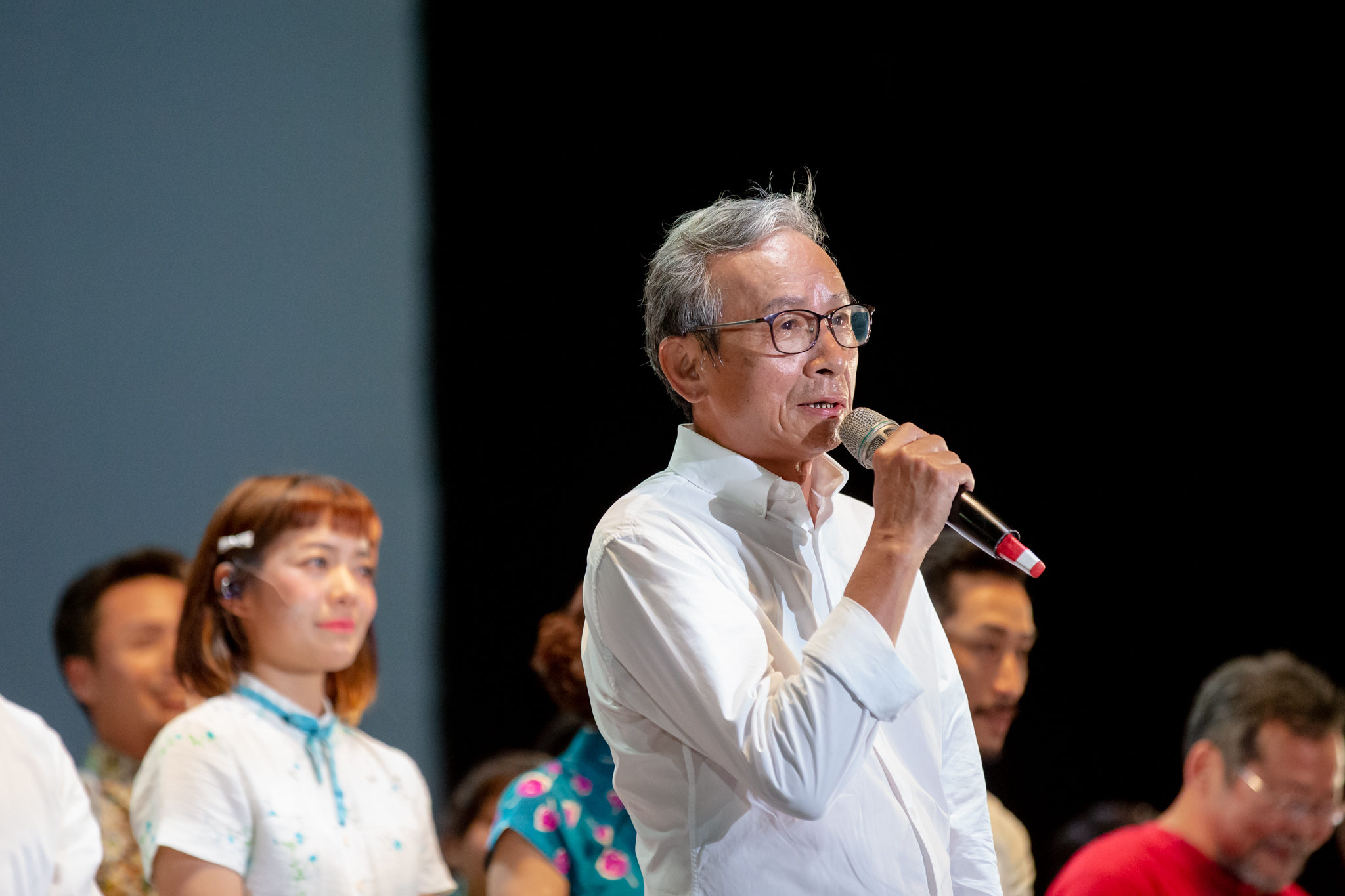 吳念真穿白襯衫、戴眼鏡，手拿麥克風在台上致詞